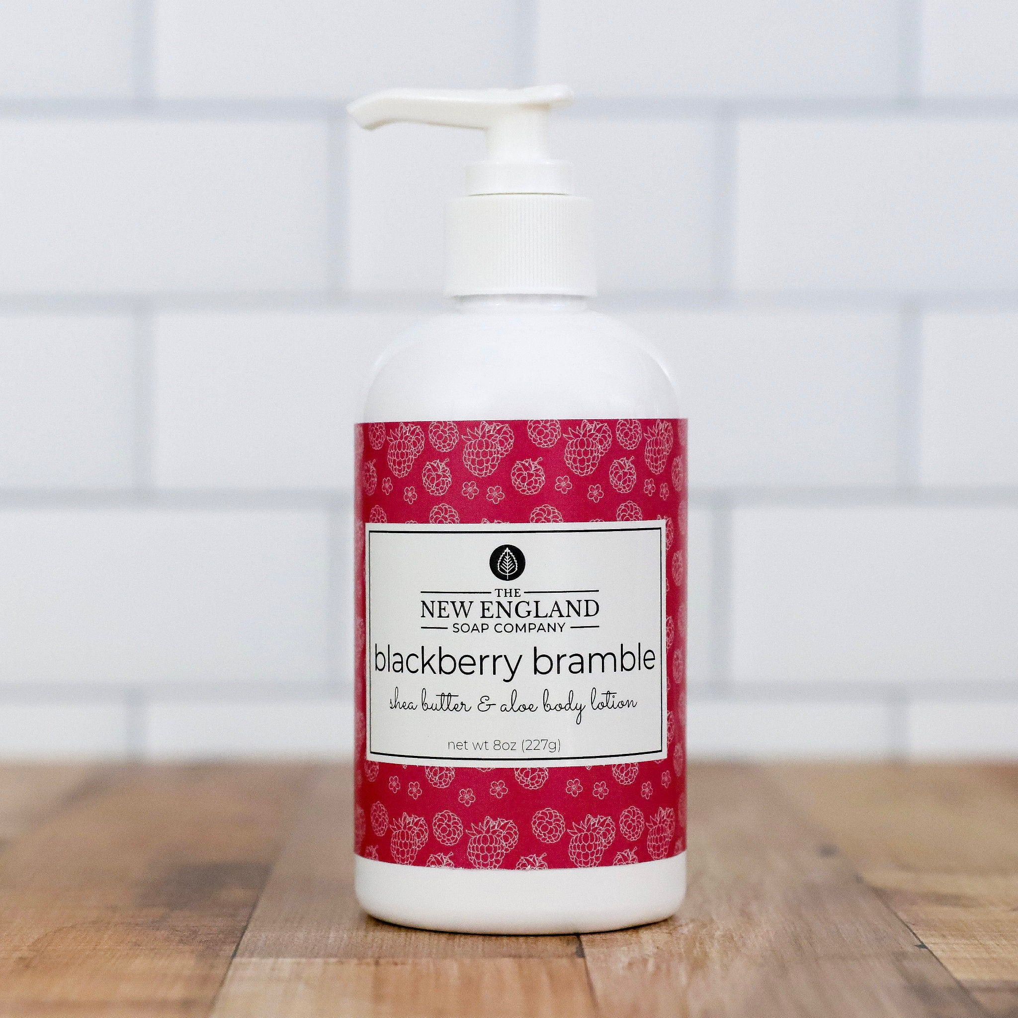 White bottle of New England Soap Company blackberry bramble shea butter & aloe body lotion on a wooden surface with a tiled wall background.