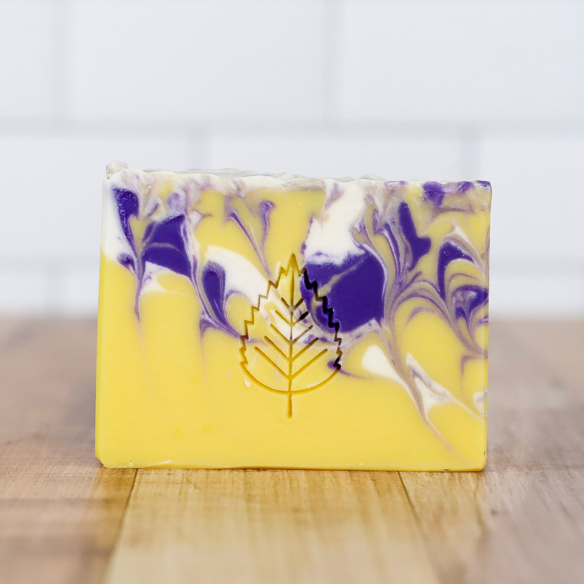 Yellow soap bar with purple and white swirls and a leaf logo on a wooden surface.