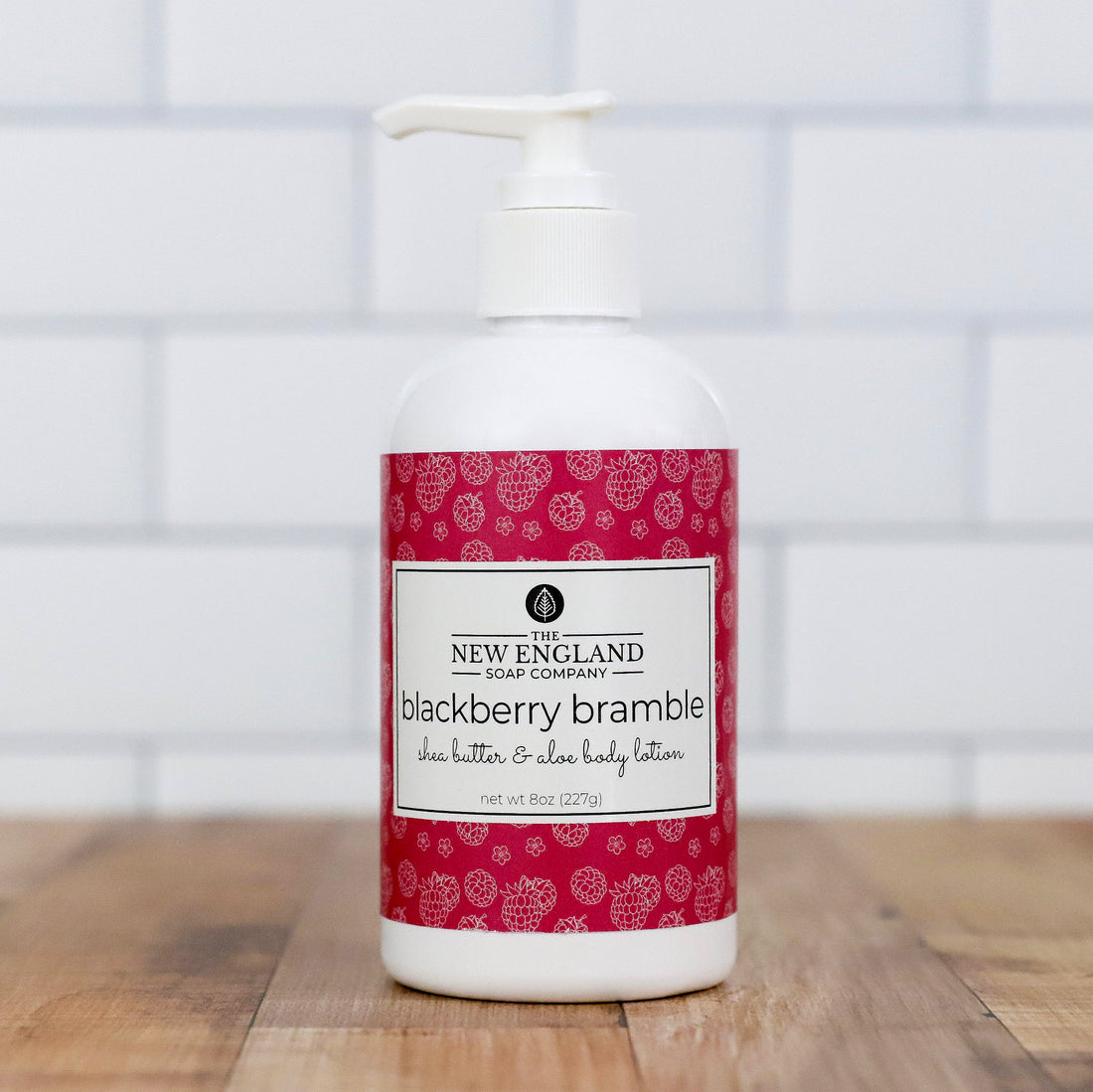 White bottle of New England Soap Company blackberry bramble shea butter &amp; aloe body lotion on a wooden surface with a tiled wall background.