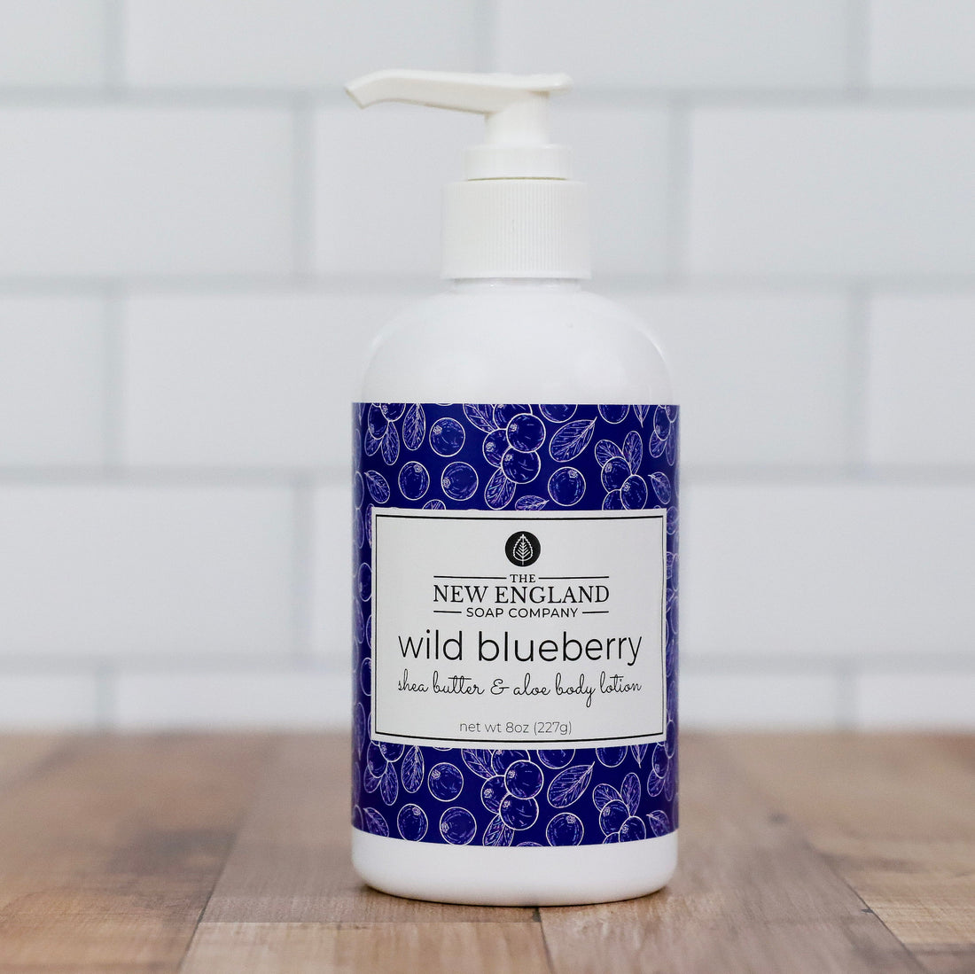 Bottle of New England Soap Company wild blueberry body lotion on a wooden surface with a white tiled wall background.